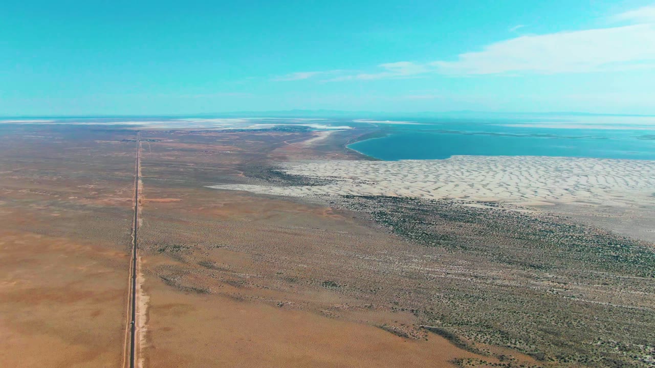 High aerial frame in Guerrero Negro, Baja California Sur, Mexico, showing a straight road entering brown desert toward pale salt flats and blue water, with no visible people.