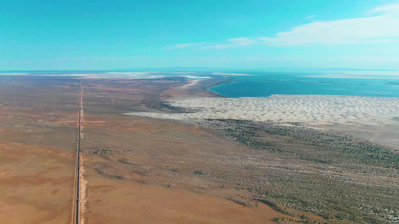Straight road through arid terrain with salt flat and water beyond.