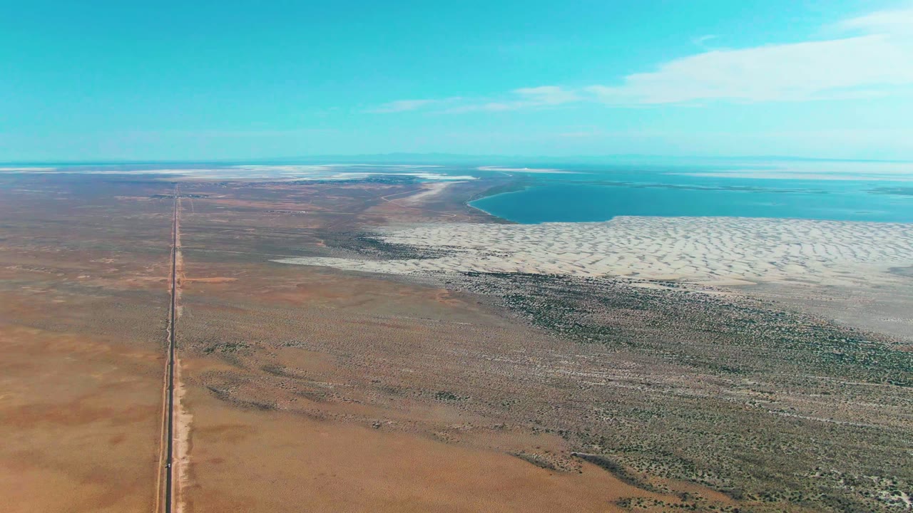 High drone-style view in Guerrero Negro, Baja California Sur, Mexico, showing the road running upward through brown desert toward white salt flats and turquoise-blue water, with no visible people.