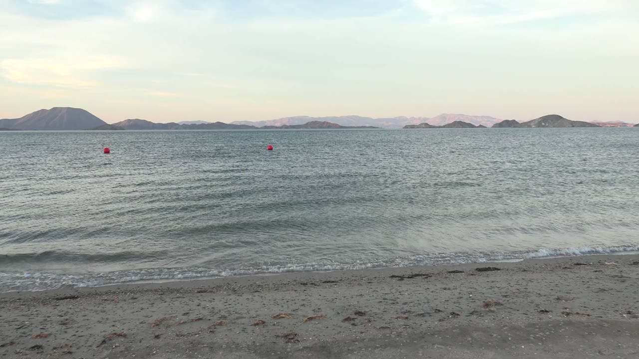 Bahia De Los Angeles shoreline at the start of the clip, with small waves reaching gray sand, several red buoys offshore, distant islands on the horizon, and no visible people.