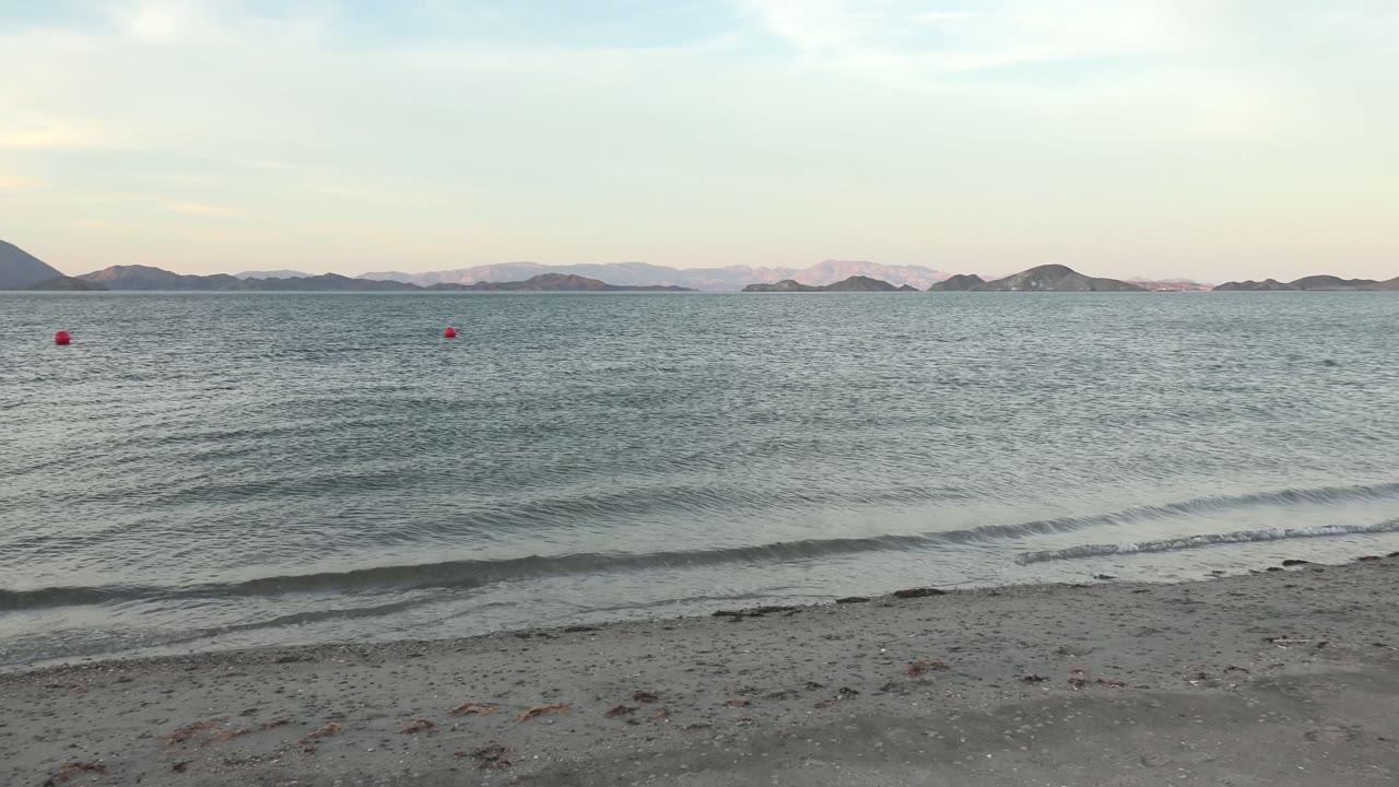 Bahia De Los Angeles water view at 0:02, with rippled gray-blue water, thin surf near the sand, red buoys in the midground, island silhouettes, and no visible people.