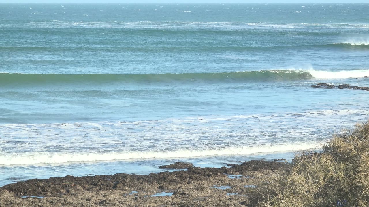 At La Bocana, Baja California Sur, a blue ocean swell forms behind dark rocky foreground ledges, with pale foam near shore and no visible people.