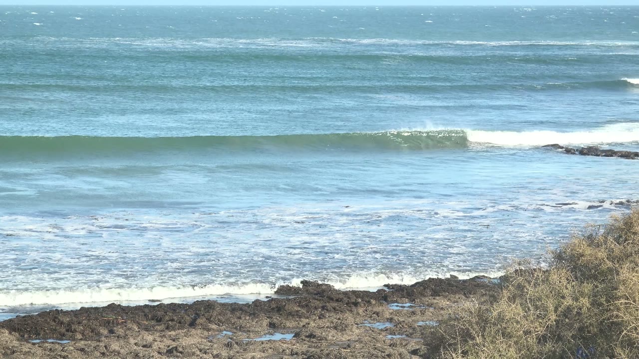 At La Bocana, Baja California Sur, white spray rises from the breaking wave while dark coastal rocks and scrub remain fixed in the lower frame, with no visible people.