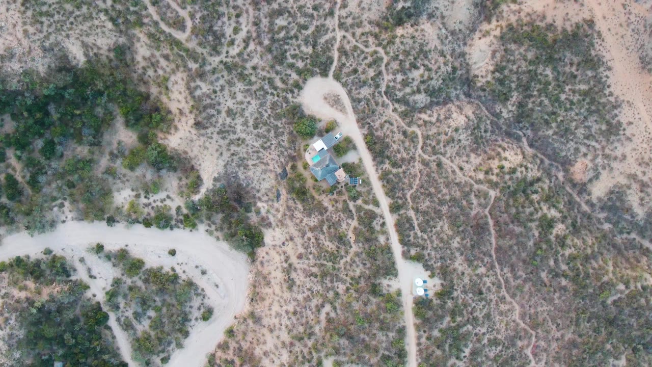 Aerial frame over Cabo Pulmo showing a curved dirt road at left, scrub-covered terrain, small light-roofed buildings near the upper right, and no visible people.