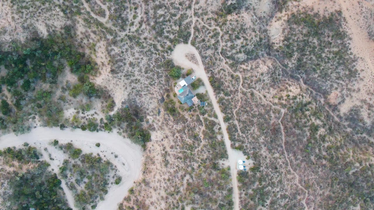 Overhead Cabo Pulmo frame with intersecting pale tracks, mottled green and tan scrub, a tiny vehicle lower on the road, small structures near the top, and no visible people.