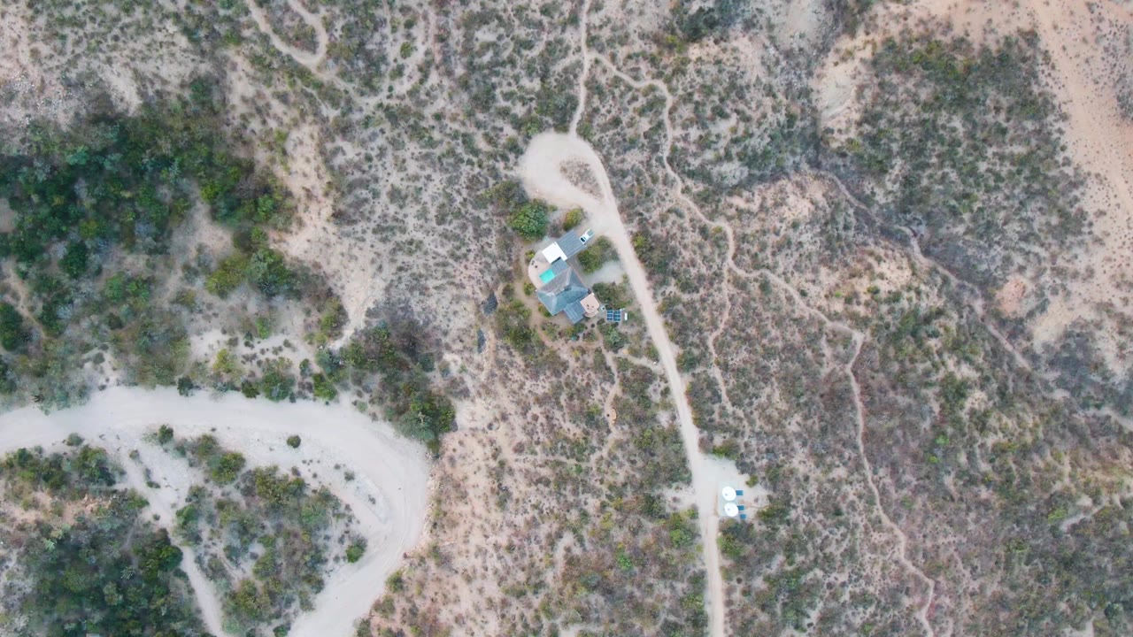 Drone view of dry scrubland with a curved dirt road and small structures beside another road.