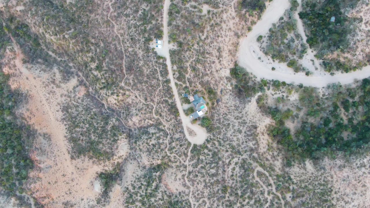 Aerial frame over Cabo Pulmo terrain with the winding road crossing scrub-covered hills and the vehicle slightly farther along the route, with no visible people.