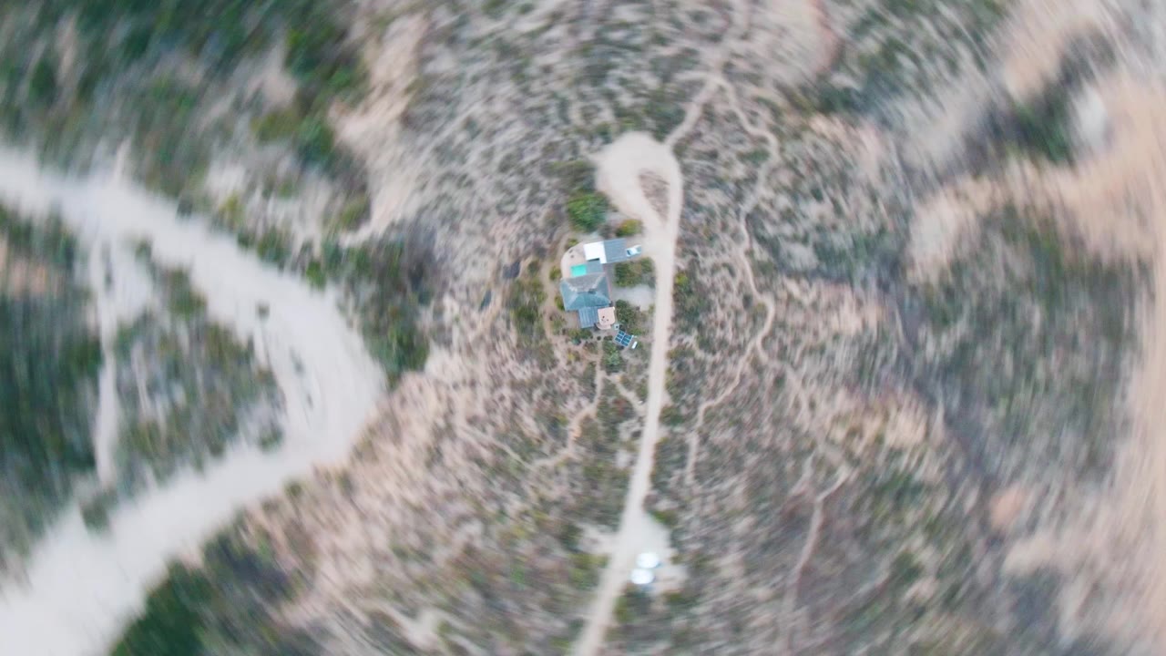 Overhead view in Cabo Pulmo, Baja California Sur, with a small cabin compound near pale dirt roads, scrub-covered desert terrain, and no visible people.