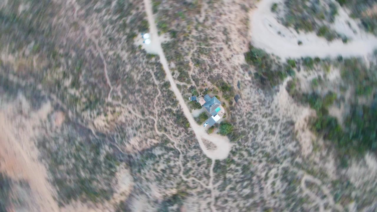 High overhead view in Cabo Pulmo with the cabin compound at the right side of frame, looping dirt access roads, sparse green scrub, and no visible people.