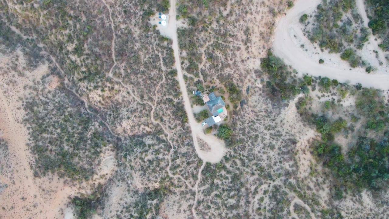 Overhead frame of a pale dirt road, a small turquoise-roofed house, scrubby Cabo Pulmo desert terrain, and a white vehicle near the upper road with no visible people.