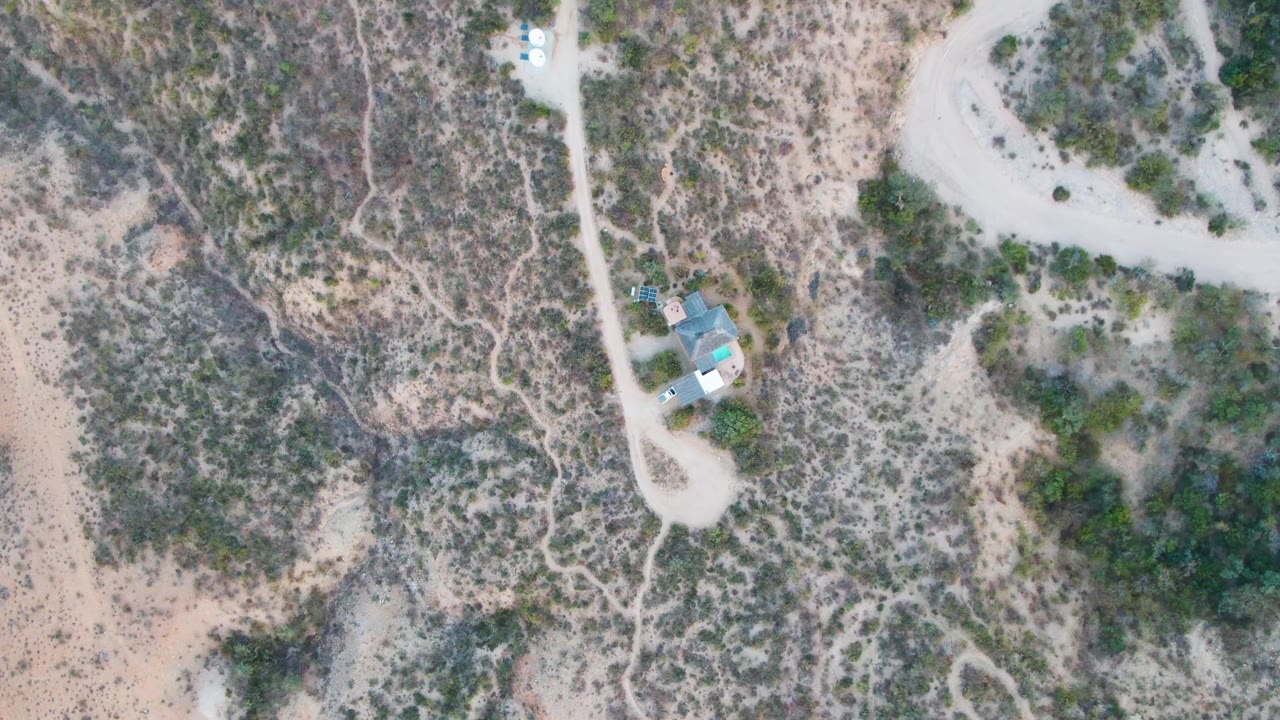Top-down Cabo Pulmo frame with a white vehicle on the dirt road near the small turquoise-roofed house, dry sandy ground, scrub vegetation, and no visible people.