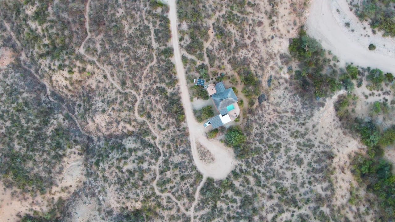 Overhead view in Cabo Pulmo, Baja California Sur, showing a pale dirt road, a small turquoise-roofed building, dry scrubland, and no visible people at the start of the clip.