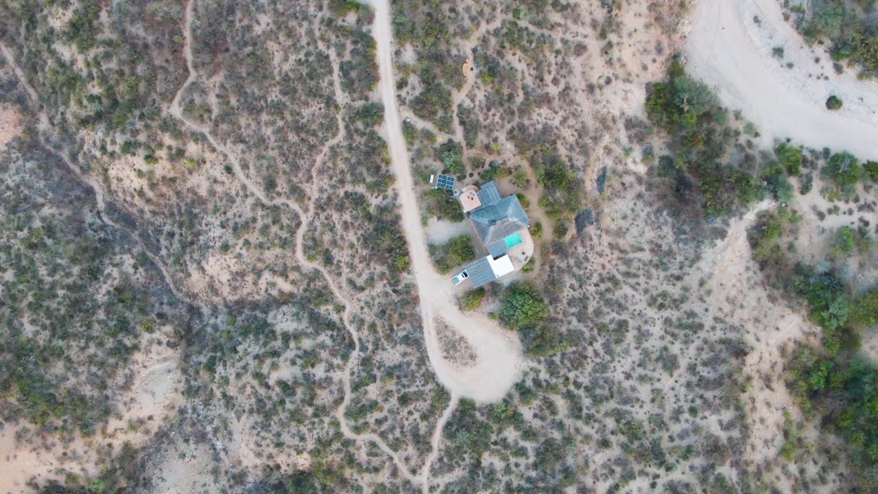 Top-down Cabo Pulmo frame showing the building and nearby light vehicle beside the dirt road, with branching dry paths through scrub and no visible people.