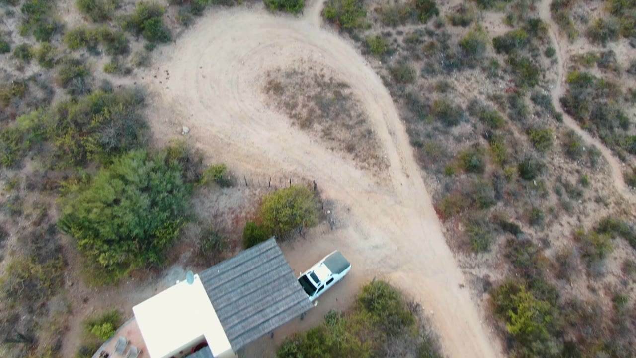 Opening overhead frame in Cabo Pulmo, Baja California Sur, showing a pale dirt loop beside a small roofed structure, a white vehicle, scrub vegetation, and no visible people.