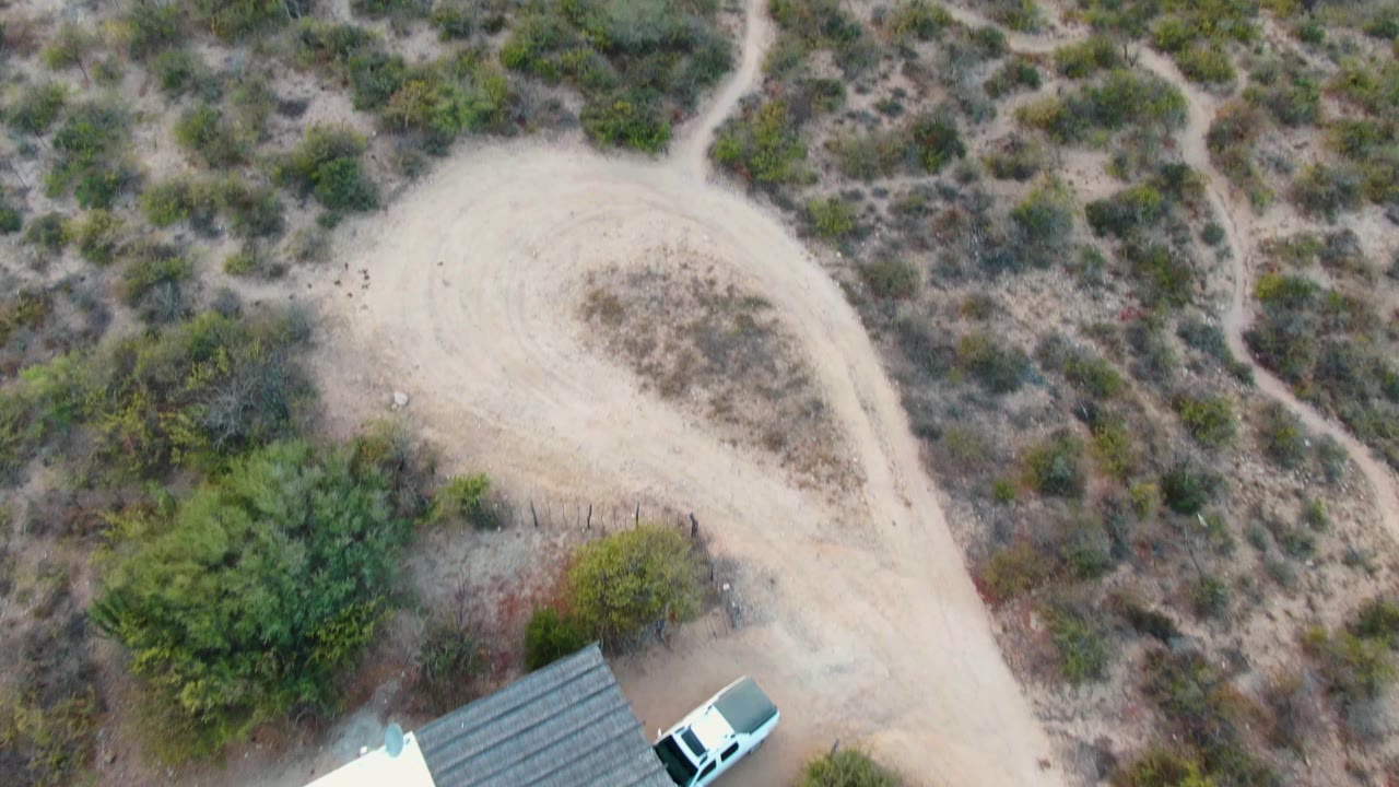 Aerial view of the house roof, white vehicle, dirt curve, and surrounding scrubland.