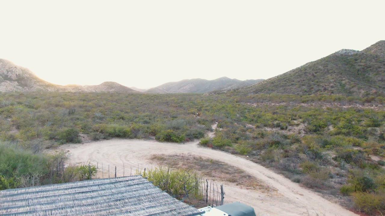Cabo Pulmo desert landscape with a roofed structure entering the lower foreground, a curving dirt road behind it, scrub vegetation, low mountains, and no visible people.
