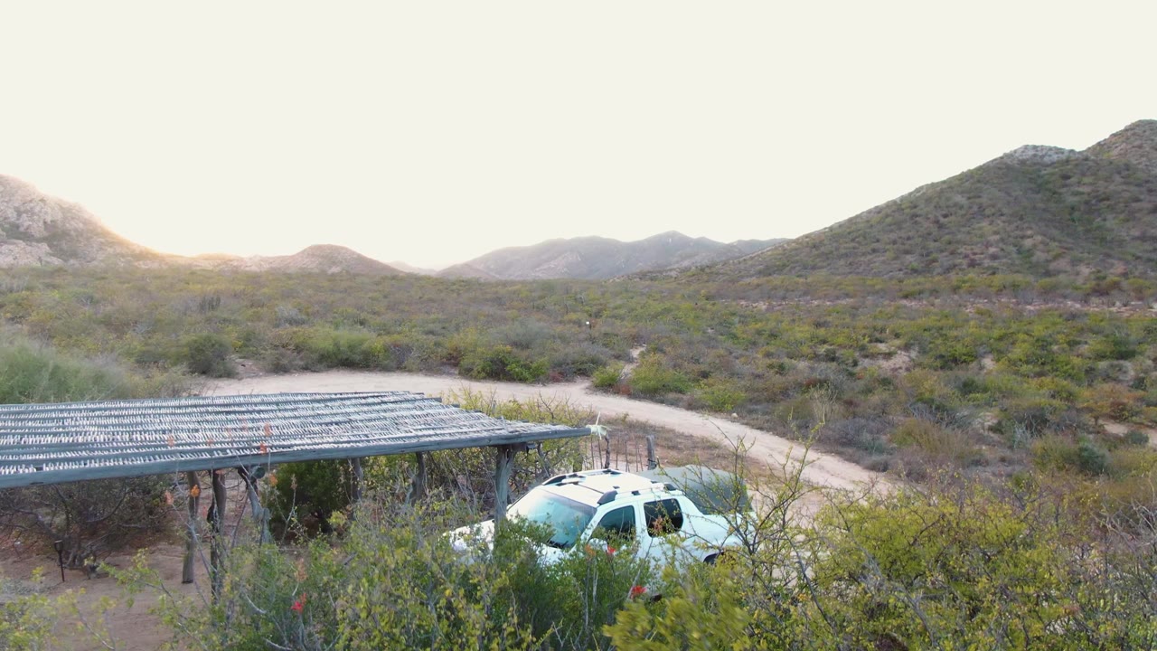 One second in, the elevated view shows the white SUV near the right side of the shade structure, framed by scrub vegetation, a pale dirt road, and Cabo Pulmo hills with no visible people.