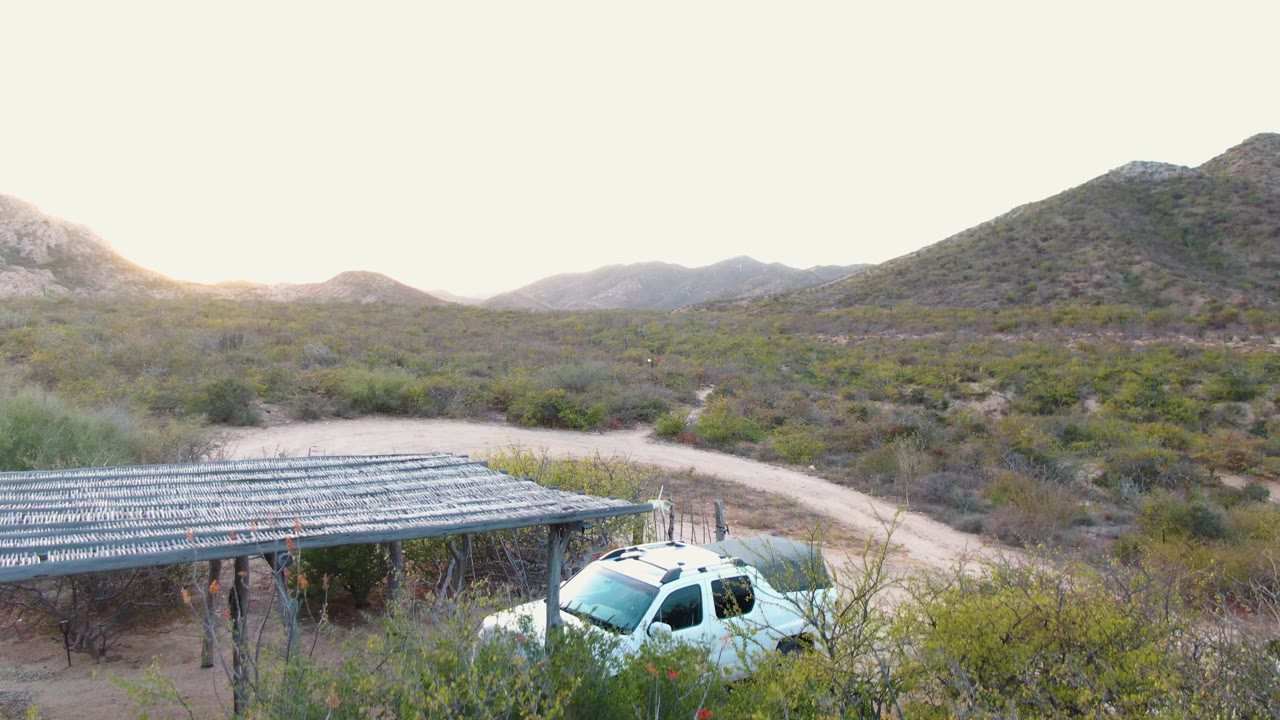 At 0:02, the SUV remains visible near the shaded roof edge, with the dirt road curving through sparse Cabo Pulmo vegetation and no visible people in the frame.