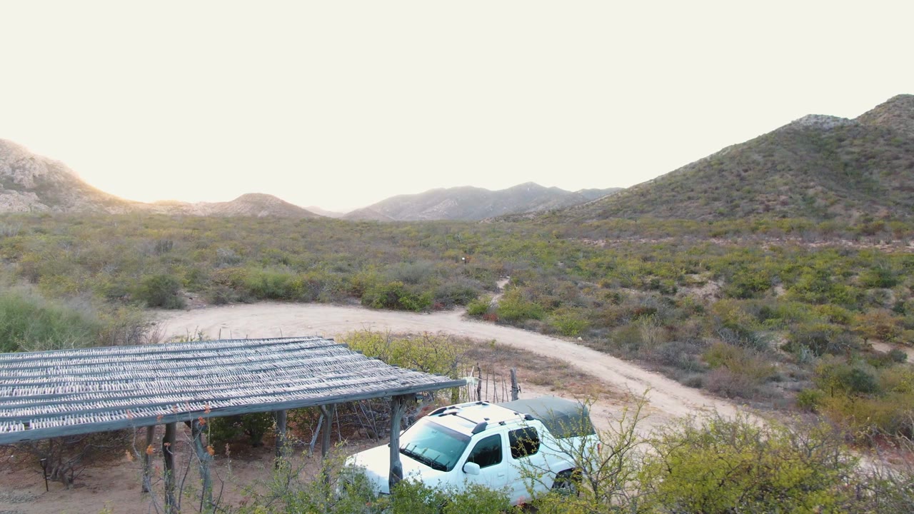 At 0:03, the white SUV sits low in the frame beside the shade structure, while the dirt road and brush-covered hills dominate the Cabo Pulmo background with no visible people.