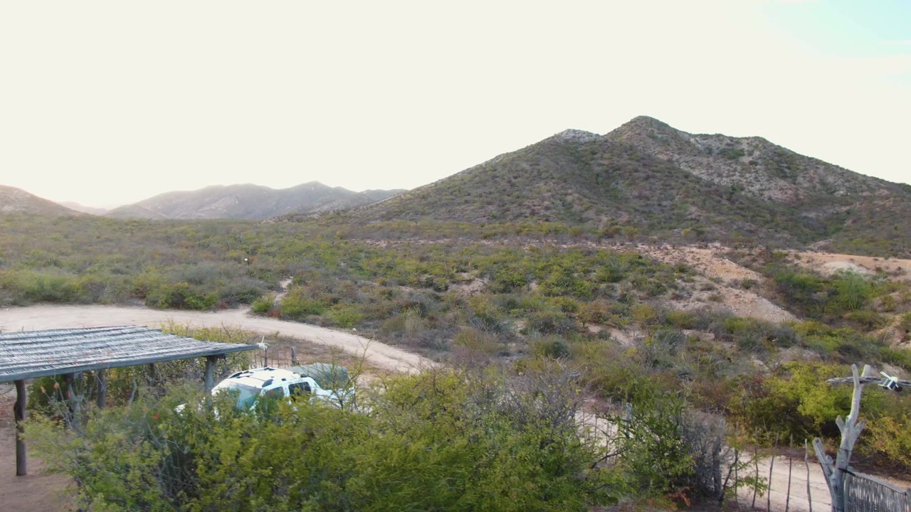 Wide elevated view in Cabo Pulmo, Baja California Sur, Mexico, showing a white car beside a metal-roof carport, desert shrubs, a dirt road, low mountains, and no visible people.