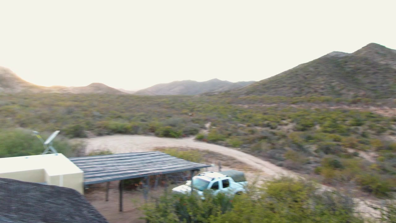Cabo Pulmo frame with the carport and white truck low in the composition, a curving pale dirt road behind them, scrub-covered slopes, and no visible people.