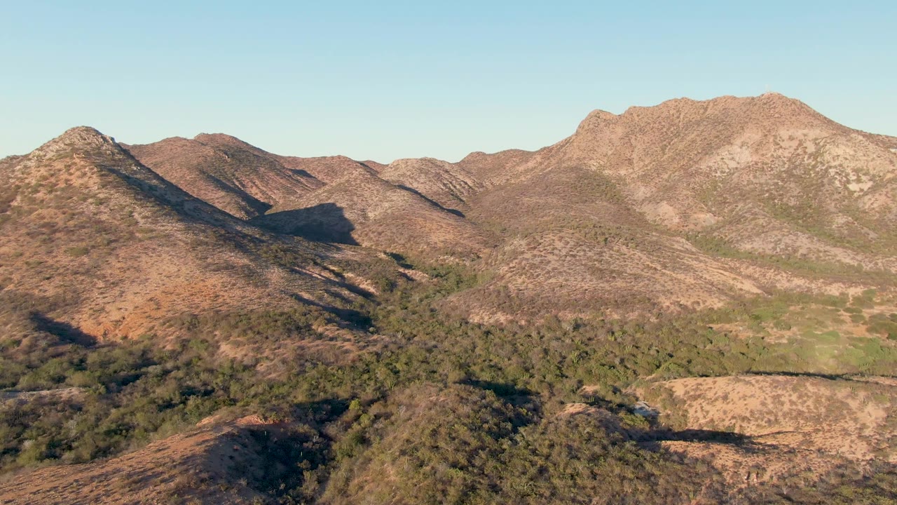Third frame in Cabo Pulmo, Baja California Sur, with the central rocky ridge more prominent, green low vegetation below it, clear sky, and no visible people.