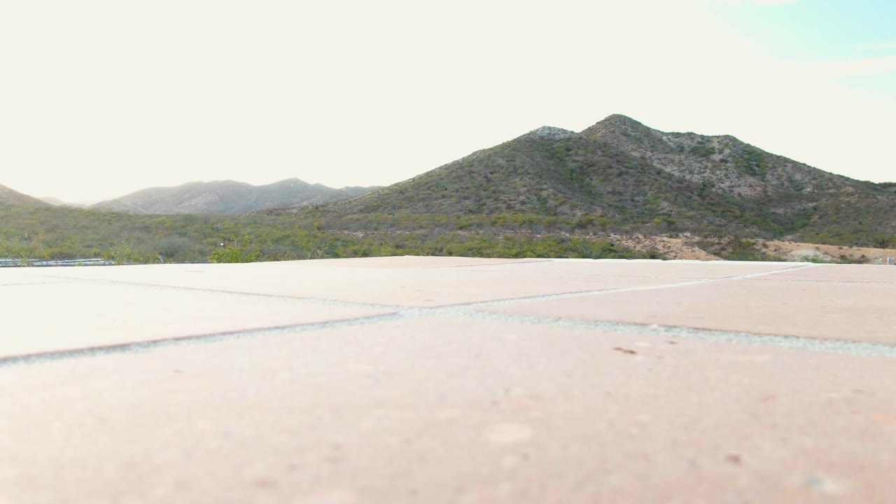 Second frame in Cabo Pulmo, Baja California Sur showing the same wide paved foreground, a narrow band of scrubland, a central rocky ridge, and no visible people.