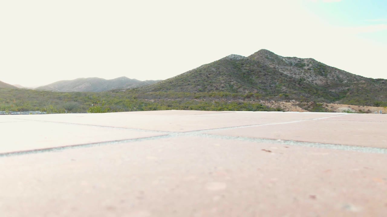 Third frame of the Cabo Pulmo clip with pale concrete or pavement across the lower frame, low green terrain in the middle distance, rocky hills behind, and no visible people.