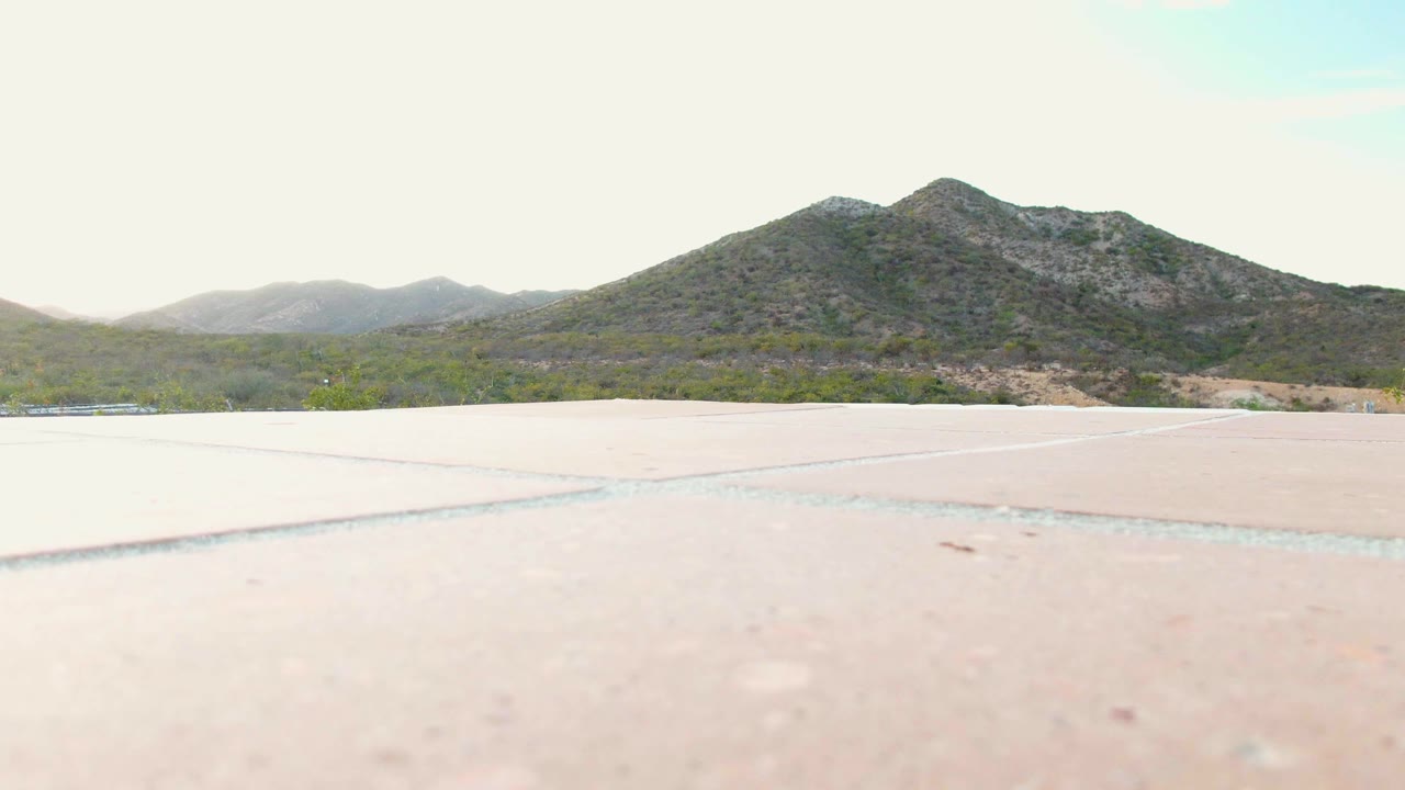Fourth frame in Cabo Pulmo, Baja California Sur where the foreground pavement remains dominant, the scrub band runs across the horizon area, rocky hills sit beyond it, and no visible people appear.