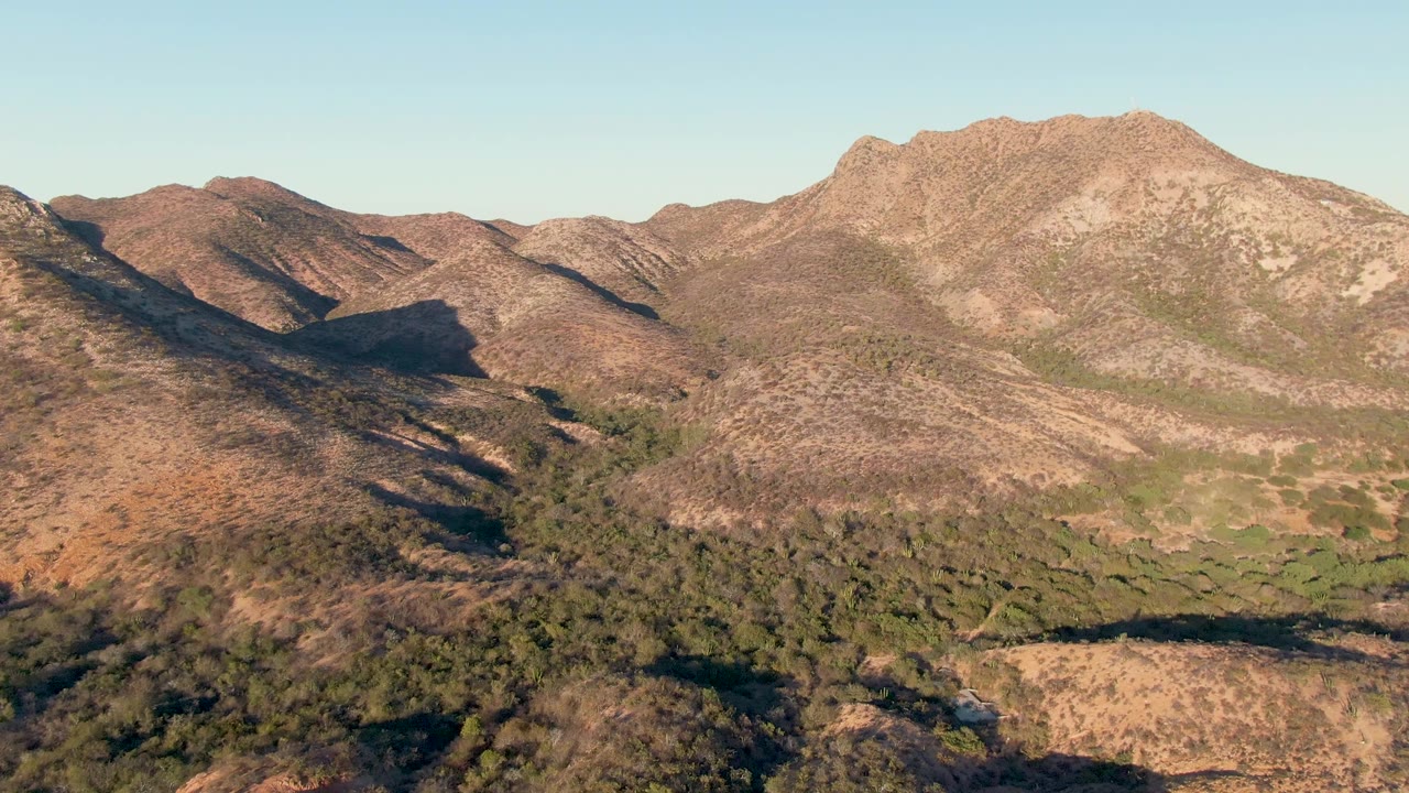Aerial view near Cabo Pulmo, Baja California Sur, showing dry brown mountain slopes, a green-brush valley, a clear pale sky, and no visible people.