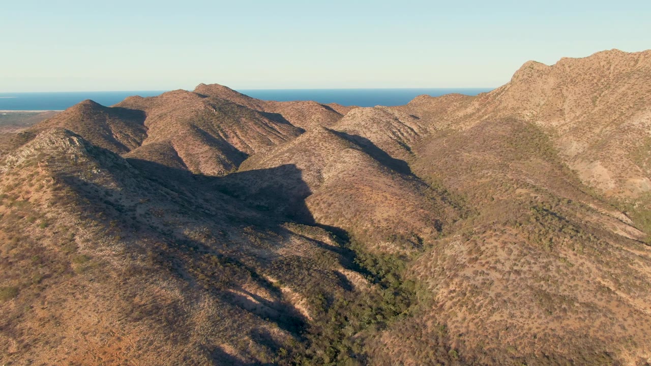 Aerial frame over brown Cabo Pulmo hills, with a dark shadowed valley in the lower left, rocky ridges across the middle, blue ocean beyond, and no visible people.