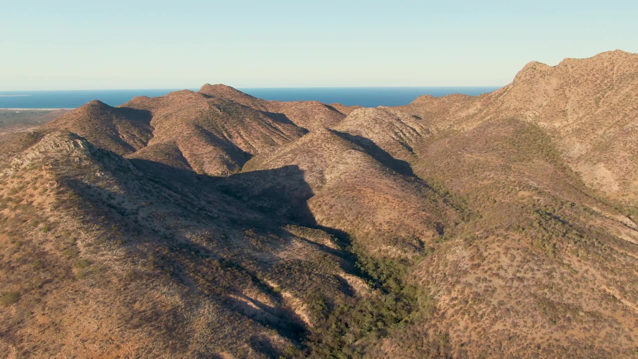 Sunlit arid hills near Cabo Pulmo stretch across the frame, with darker ravines between ridges, a thin blue ocean horizon in the distance, and no visible people.