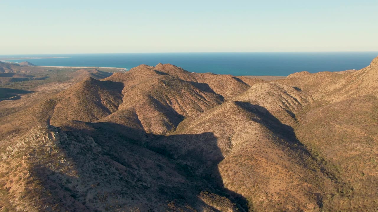 Aerial view across sunlit Cabo Pulmo desert mountains, with shadowed gullies in the center, ocean across the top of frame, and no visible people.