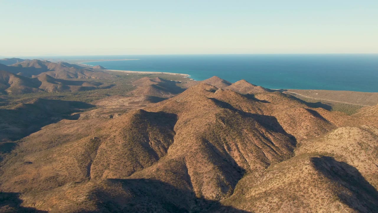 Aerial frame over Cabo Pulmo with dry ridges and valley shadows across the foreground, a strip of coastline in the distance, blue ocean, and no visible people.