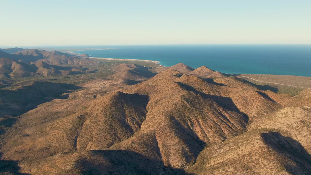 Aerial frame in Cabo Pulmo showing rugged brown hills across the lower frame, layered coastal mountains to the left, open blue ocean behind them, and no visible people.