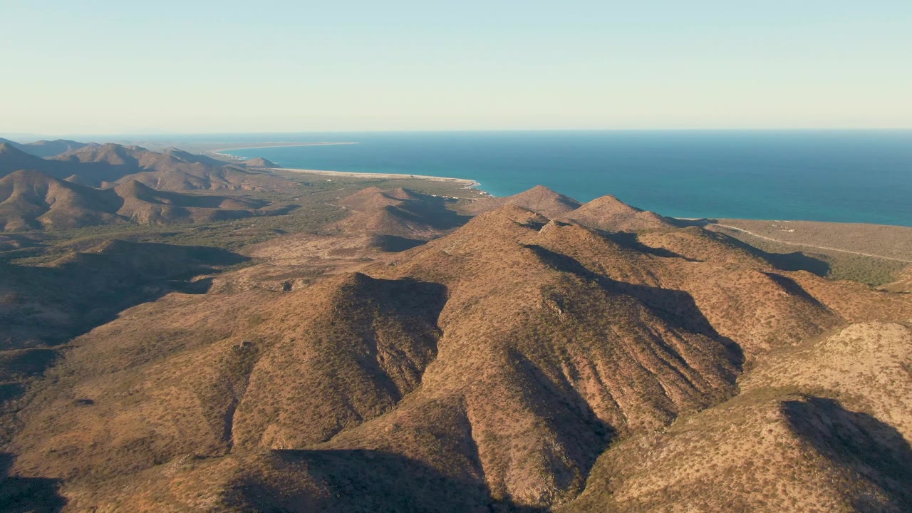 Aerial frame over Cabo Pulmo with the main ridge cluster slightly lower in frame, broad dry hills spreading left, blue ocean across the background, and no visible people.
