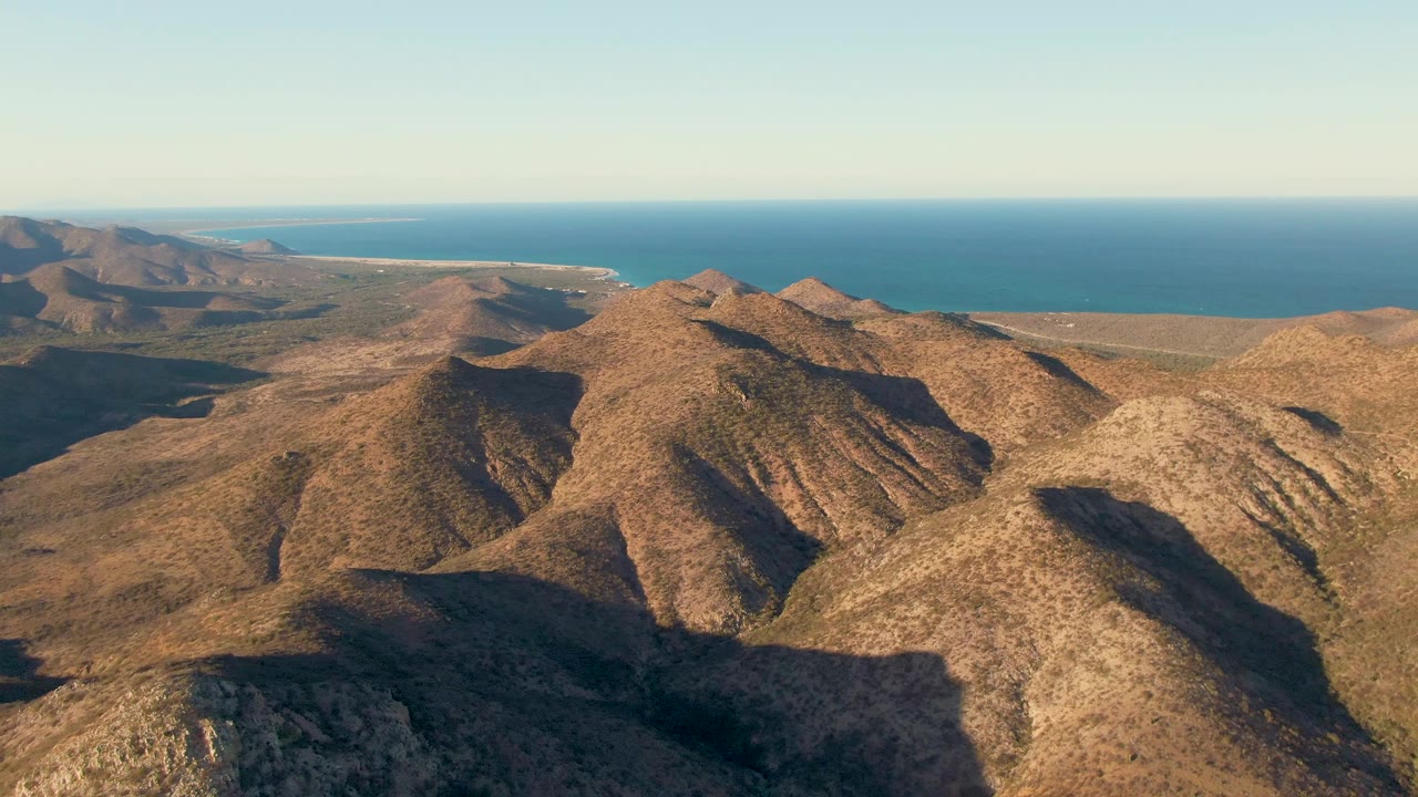 Aerial view over dry brown mountain ridges in Cabo Pulmo, Baja California Sur, with blue ocean beyond and no visible people.