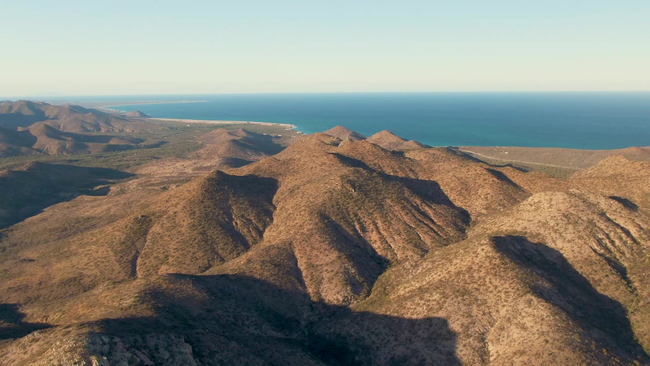 Aerial view of layered brown mountain slopes in Cabo Pulmo with the coastline and open ocean behind them, with no visible people.
