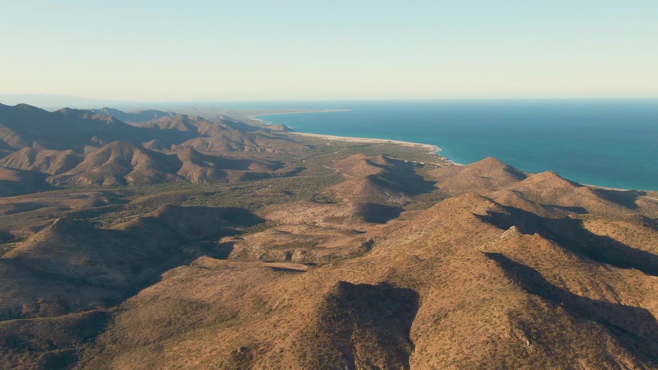 Cabo Pulmo aerial frame showing sunlit brown hills across the foreground, darker mountains on the left, a bright sandy coast on the right, blue ocean, and no visible people.