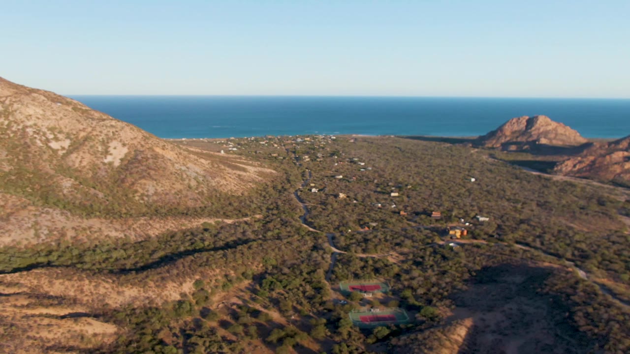 Aerial frame of Cabo Pulmo hills with a sloping foreground ridge, winding dirt roads, scattered buildings, rocky headlands, ocean, and no visible people.