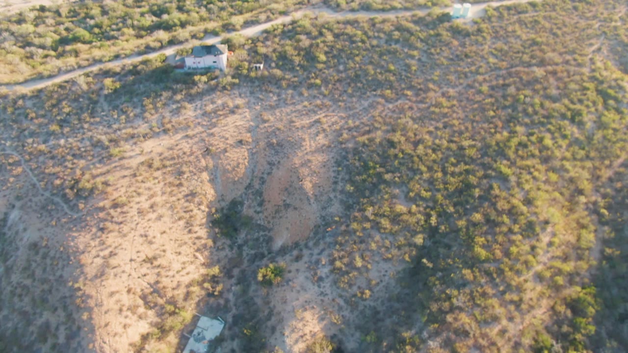 Aerial frame over Cabo Pulmo, Baja California Sur, showing a sandy dry hillside crossed by a dirt road, a small light-colored building near the upper edge, sparse bushes, and no visible people.