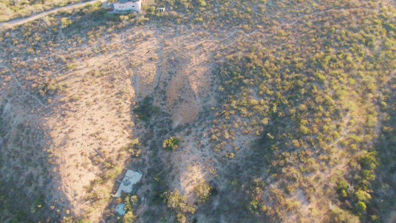 Top-down aerial of sandy hillside with brush, shadows, and a trailer near the lower edge.