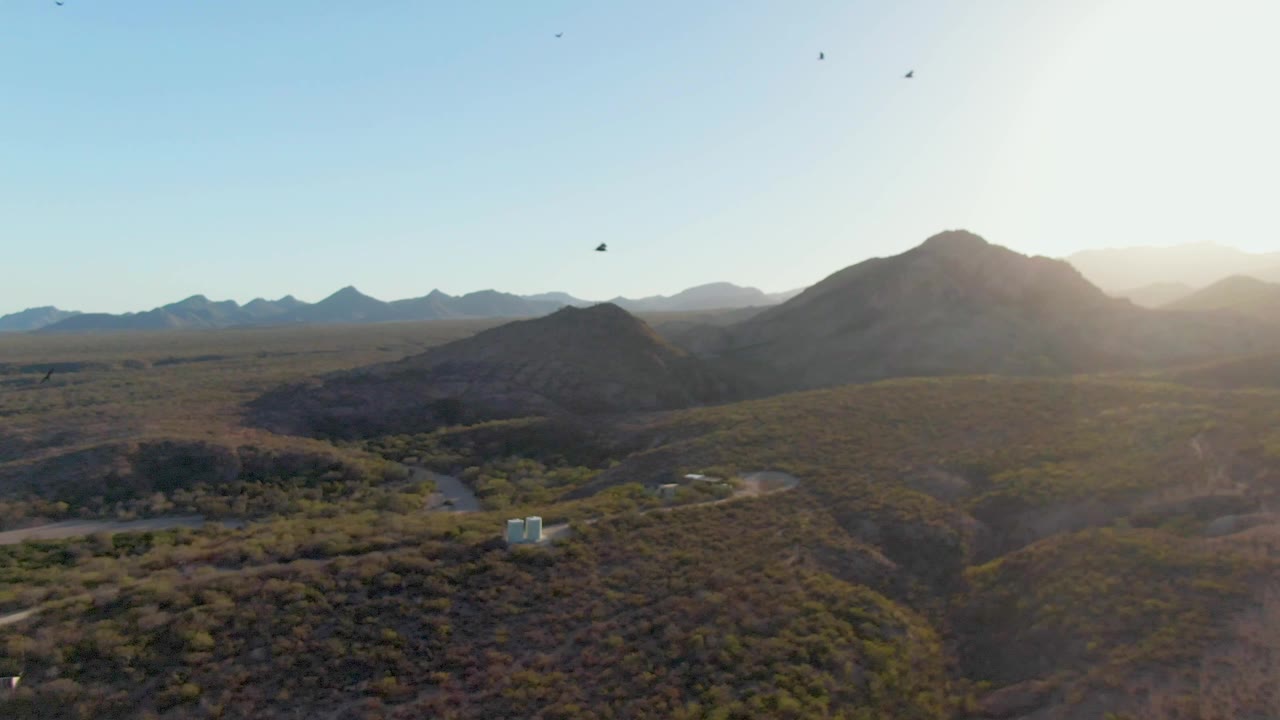 Birds pass through a clear sky over Cabo Pulmo hills as bright sun washes the upper right of the frame, with desert roads and no visible people below.
