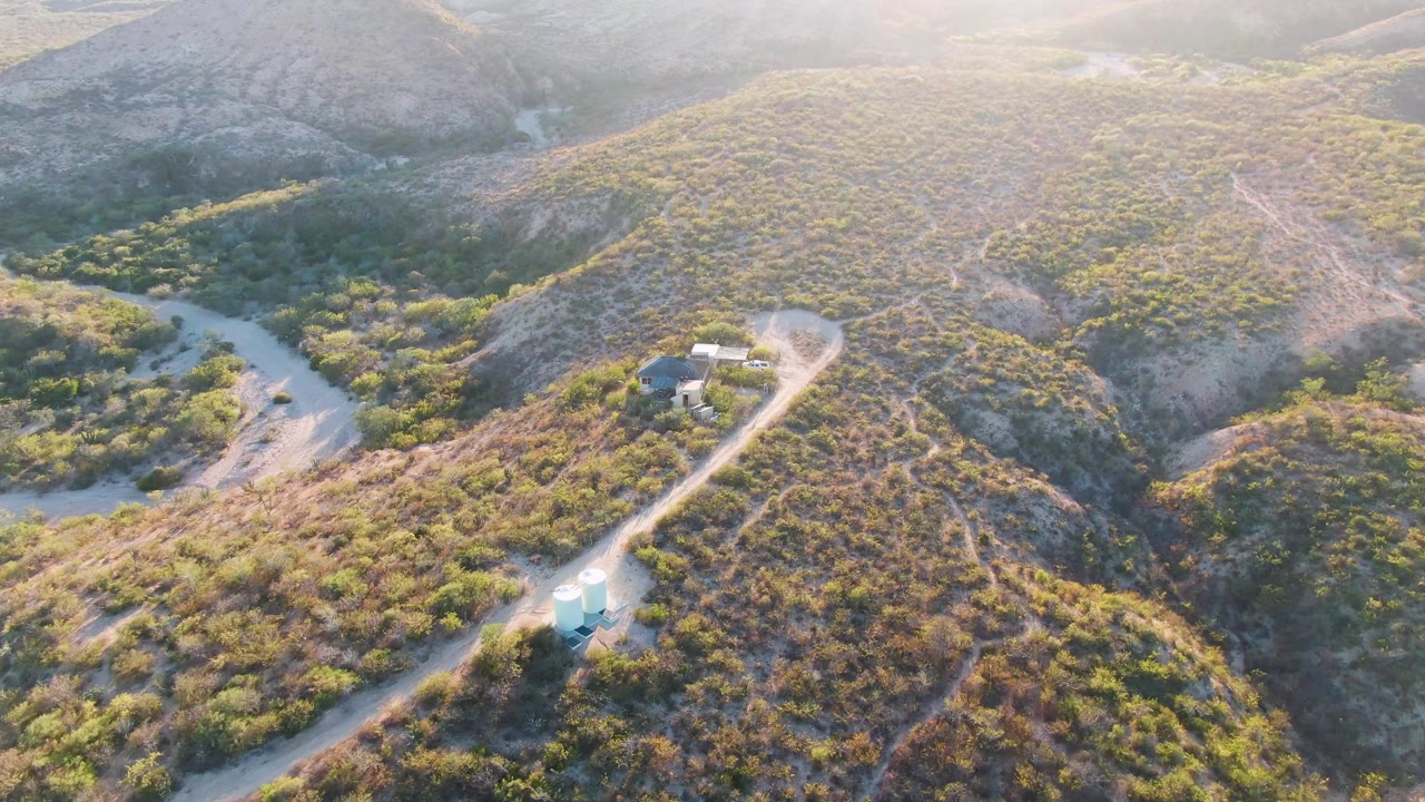 Aerial frame in Cabo Pulmo, Baja California Sur, with the road sweeping from the lower edge toward a hillside building area, the blue car on the route, and no visible people.
