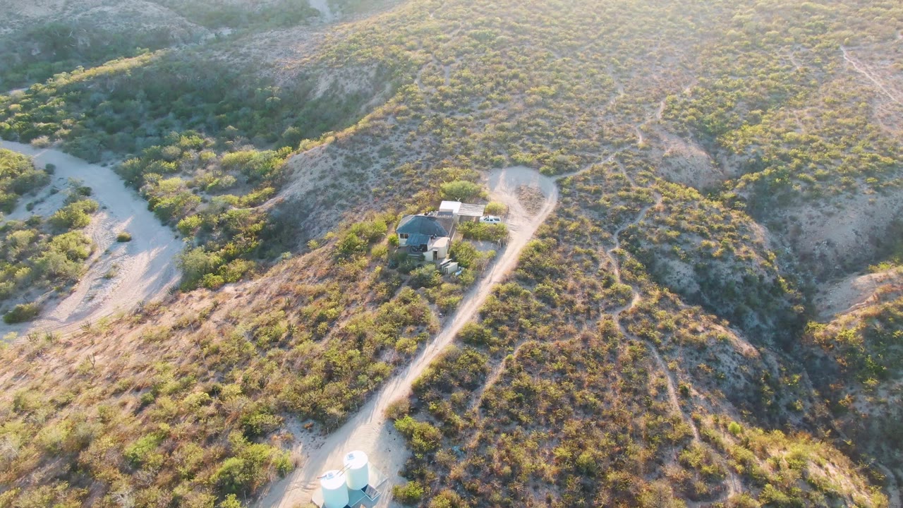 Drone view over dry hills, a curving road, and small structures near the ridge.