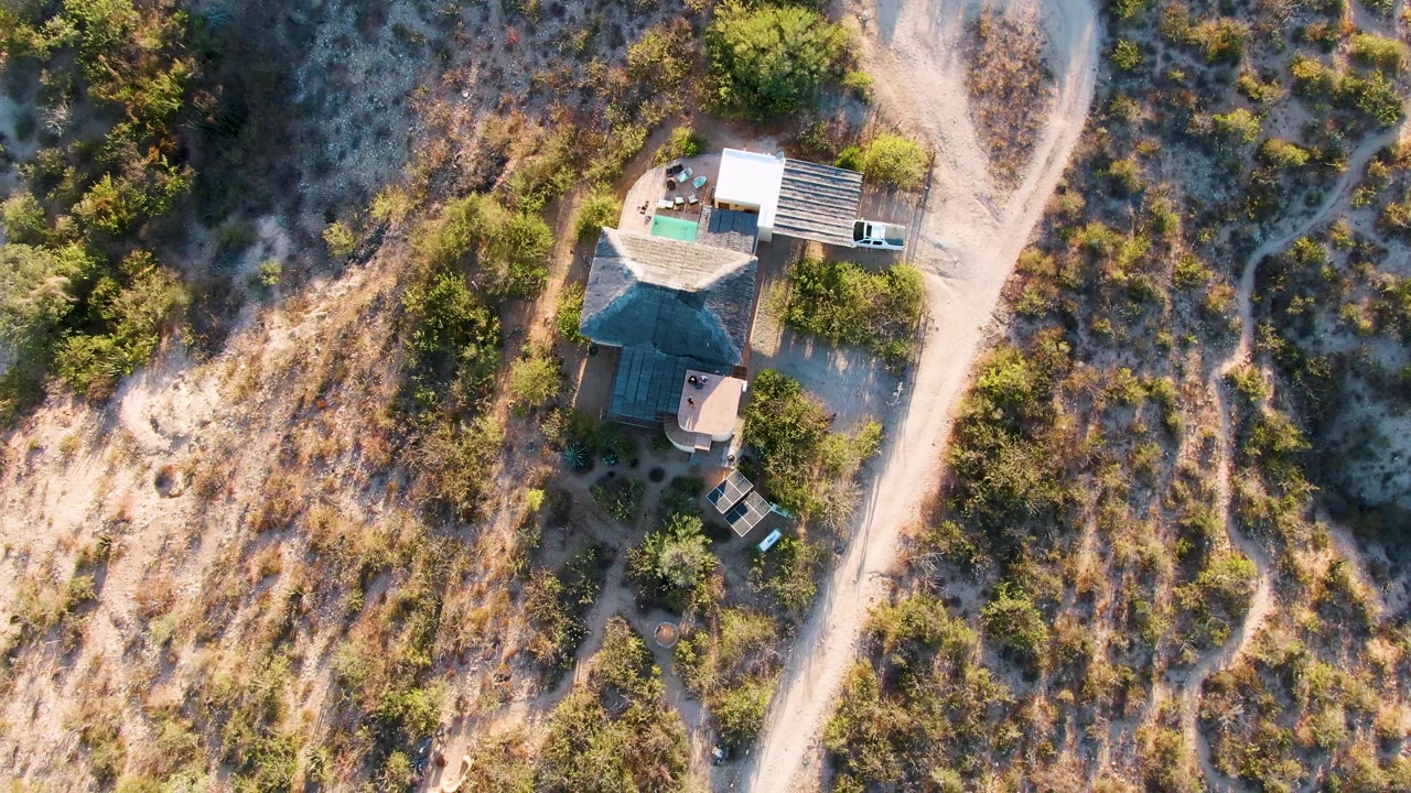 High aerial frame in Cabo Pulmo, Baja California Sur showing a small house beside a pale dirt driveway, scattered shrubs, dry grass, and no visible people.