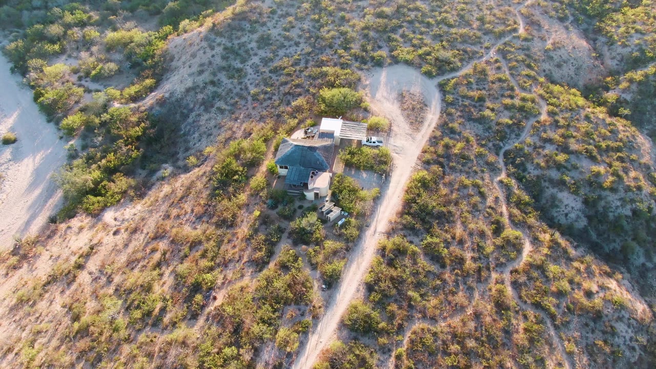Aerial frame over Cabo Pulmo, Baja California Sur, showing a small hillside house beside a pale dirt road, surrounded by dry brush and green shrubs, with no visible people.