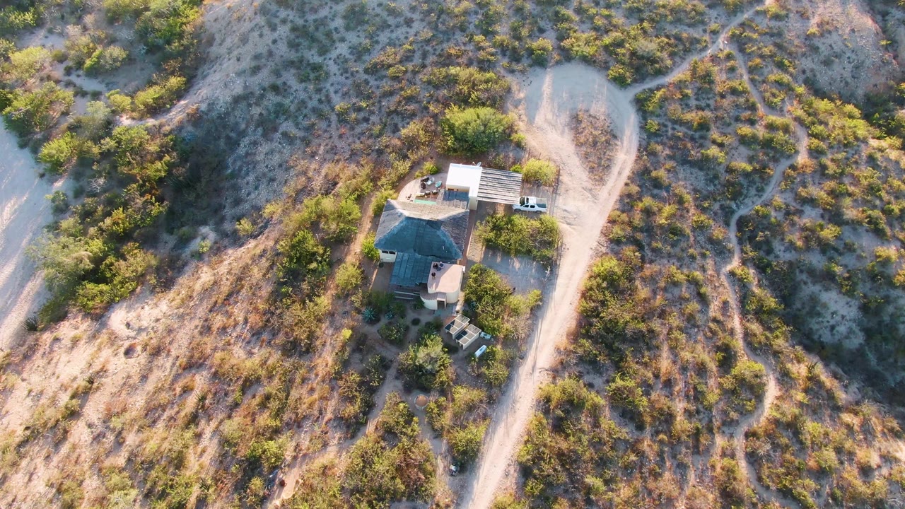 Overhead aerial in Cabo Pulmo, Baja California Sur, showing the house and nearby dirt road more centered in the frame, with dry brown terrain, green vegetation, and no visible people.