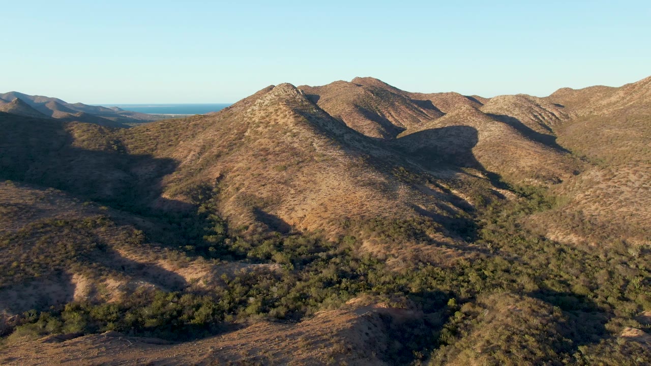 Cabo Pulmo aerial view at the start of the clip, with dry brown ridges, a green valley floor, distant ocean at left, clear sky, and no visible people.