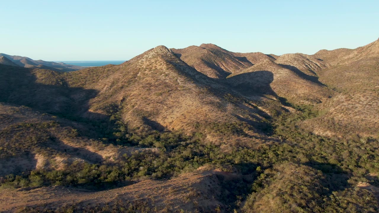 Mid-early frame of the Cabo Pulmo mountain view, showing the rounded central ridge, green vegetation below, distant blue ocean to the left, and no visible people.